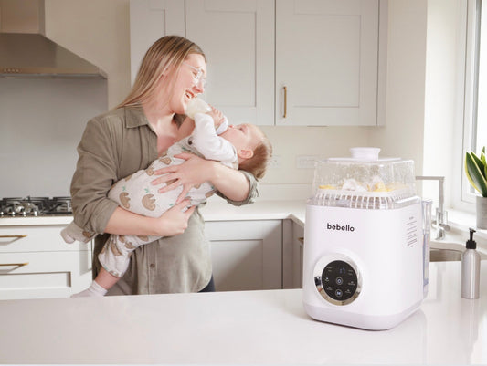 Mother feeding baby with a bottle in the kitchen standing next to Bebello Washer on the counter